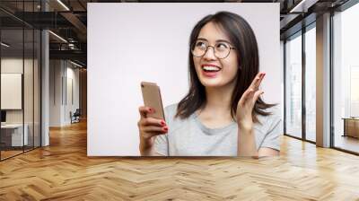 Joyful charming asian woman in spectacles with long dark hair holding modern smartphone isolated over white background in studio Wall mural