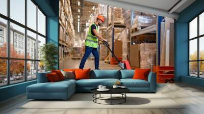 male staff worker in warehouse uses hand pallet stacker to transport goods, alone, dressed in working clothes and safety hard hat.Skilled warehouse employee pushing manual pallet jack Wall mural