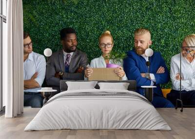 new female worker sits with others on chair, waiting for interview, holding box with things for working. new diverse colleagues look at her with contempts Wall mural