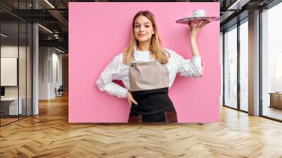 nice woman waitress in apron, offering cup of delicious tasty coffee on tray, stand smiling, friendly staff of restaurant. isolated over pink studio background Wall mural