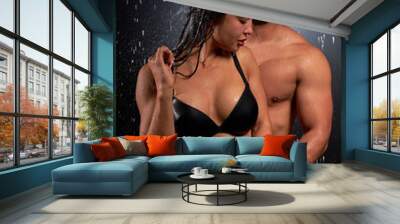 Portrait of lovely beautiful couple with athletic body under heavy rain on black background. sexy wet caucasian man kisses hugging woman while standing behind her. Love, passion concept Wall mural