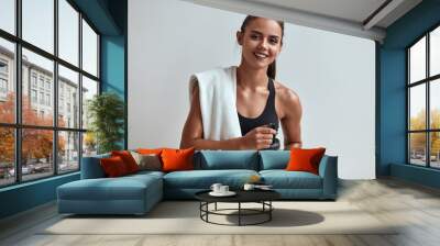 Positive smiling attractive woman in sportswear holding water bottle with towel on shoulders. Isolated in grey studio, room Wall mural