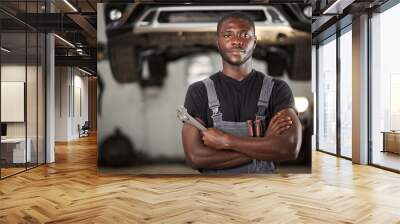 professional black auto mechanic looking at camera, handsome black guy in uniform is keen on repairing car. automobile in the background Wall mural