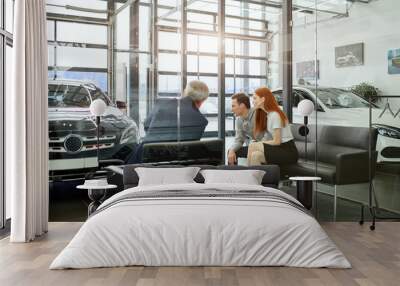 Sales agent at the table desk telling about car features to cheerful couple of customers at car dealership office Wall mural