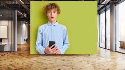 schoolboy is in shock by news on phone, stand with open mouth and big eyes looking at camera. isolated over green background Wall mural