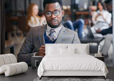 Sedate black man in grey tuxedo and eyeglasses sit looking at camera while his colleagues sit with smartphones behind him Wall mural