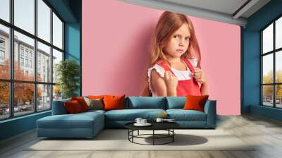 Side view on upset caucasian girl wearing red overalls, having lon hair stand isolated over pink background. Looking at camera Wall mural