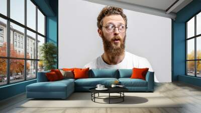 surprised caucasian man with curly hair and beard stares in amazement aside, dressed in casual white Wall mural