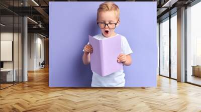 Sweet boy wearing white t-shirt and glasses for eyes reading book with purple cover, stand in shock after reading, bright emotions of kid boy. Portrait of wise child isoated over purple background Wall mural