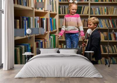 two school children help each other to get a book from shelf, stand talking, in library Wall mural