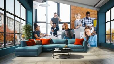 young Caucasian handsome guy with cell phone is standing behind his working partners in the modern office with panorama window Wall mural