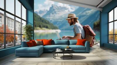 Young man freelance traveler wearing a hat and typing on his laptop, surrounded by breathtaking scenery Wall mural