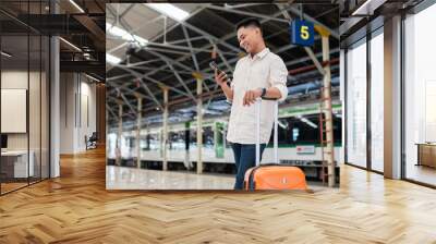 A smiling young man stands with his suitcase at a train station, checking his smartphone. He appears content and ready for travel or business trip, surrounded by the busy station environment. Wall mural