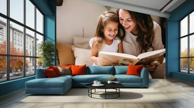 Preschool age girl laughs happily while sitting with her mom reading a story book Wall mural