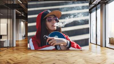 Vaping girl. Young woman with skateboard vape e-cig. Pretty young female in black hat, red clothing vape ecig, vaping device at the sunset. Toned image. Hip-hop style. Wall mural