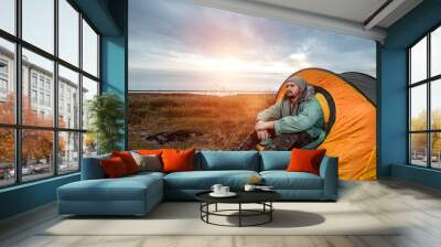 A bearded man sits camping in a tent against the backdrop of nature and the lake. The concept of travel, tourism, camping. Wall mural