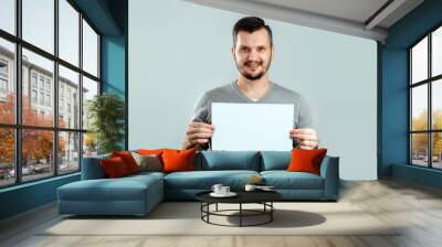 A young, attractive man holding a blank white A4 sheet, on a light background. mockup, layout, copy space. Wall mural