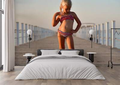 Child girl in a swimsuit stands on the pier at sunset Wall mural