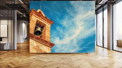 A bell tower with a bell hanging from it. The sky is blue and there are clouds in the sky Wall mural