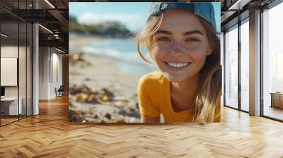 A smiling woman wearing a baseball cap sits on a sandy beach with ocean waves behind her. Wall mural