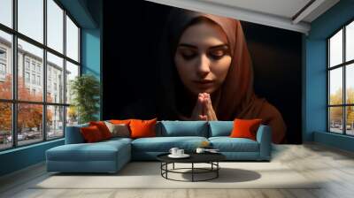 Beautiful young woman in hijab is praying with closed eyes. Muslim female close up in traditional clothes with dark background. Wall mural