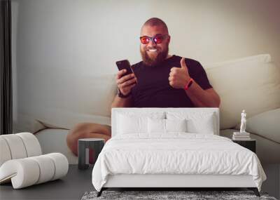 Man with blue light blocking eye glasses (yellow amber lens) smiling and make thumb up with one hand and hold his smartphone with other Wall mural