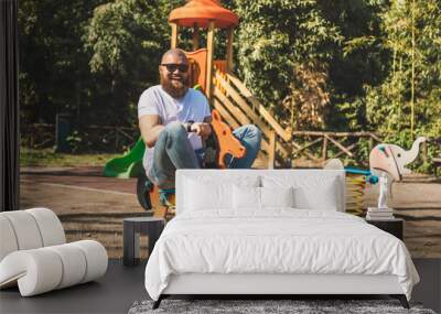 Playful crazy man (dad) riding wooden rocking horse in a park - adult guy (hipster with beard) having fun on a playground Wall mural