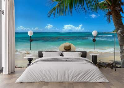 A young woman in a hat rests on a sun lounger near the clear ocean. Rear view of a woman relaxing on a snow-white beach. Vacation concept. Wall mural