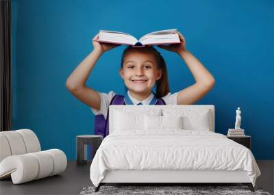 Little girl holding an open book above her head and smiling, schoolgirl concept with backpack on blue background Wall mural