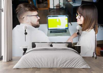 Two young coworkers dressed casual using laptop in white interior Wall mural