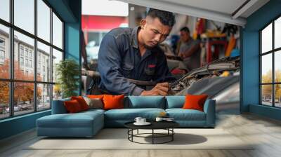 Auto mechanic working on a car engine in repair shop. Mid adult mechanic repairing a car in auto repair shop. There are people in the background. Wall mural