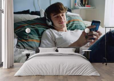 Portrait of a Young Cheerful Man Lying on His Bed Listening to Music and Using His Smart Phone Portrait of a handsome young man lying on the bed in his bedroom. He is wearing Bluetooth headphones and  Wall mural