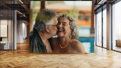 Senior woman standing on swimming pool and laughing when being kissed by her husband Elderly couple in love kissing on their romantic weekend on the swimming pool and spa Wall mural