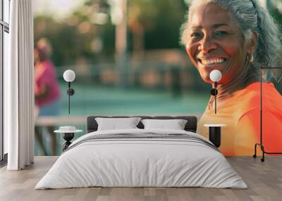 Senior black woman with orange shirt playing pickleball smiling. Happy old african american retired woman playing tennis with a friend. Representation of elderly people in the fitness industry. Wall mural