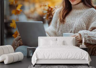 Beautiful woman working on a laptop computer while sitting in a wicker chair at a outdoors autumn cafe with cozy fall street atmosphere , a coffee cup near her hand Wall mural