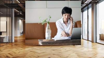 Asian self employed woman sitting on couch  talking on phone & working on laptop at home Wall mural