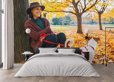 Young woman with cute dog sitting under tree in autumn park Wall mural