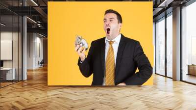 Portrait of a handsome businessman holding clock feeling sleepy and tired on yellow background. Wall mural