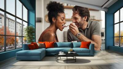 Romantic, happy and interracial couple eating a healthy yogurt together in a cute, sweet and fun kitchen romance. Loving, in love and excited husband feeding his beautiful afro wife delicious dessert Wall mural