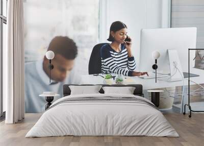 That one call confirming everything is like music to your ears. Shot of a young businesswoman taking a phone call at her office desk with a colleague working in the foreground. Wall mural