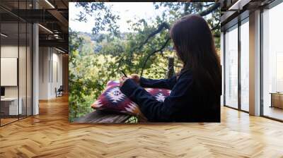 Young brown-haired woman checking messages on her cell phone on a quiet autumn morning on the terrace of her house surrounded by nature, leaning on a cushion. Wall mural