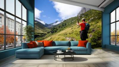 Young man taking a picture with his mobile phone on the mountain while walking on a trail dressed in ideal trekking outfit on a sunny day with blue sky and some clouds. Wall mural