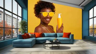 Beautiful young African-American woman in swimsuit with bottle of sunscreen cream on yellow background Wall mural