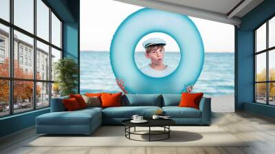 Surprised boy in inflatable swimming ring on the seashore. Portrait of a boy in the center of a blue swimming circle on the beach. Child sea travel. Boy in a sailor hat is preparing for adventures Wall mural