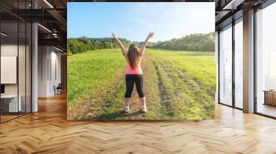 Young woman with arms up. Plus size girl is welcomes spring on the background way along the green meadow. Outdoor sports, fitness. Hello springtime. Freedom, self-confidence. Life is Beautiful. Glare Wall mural