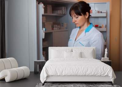 A young female doctor in white uniform is standing in a room for medical staff near a cabinet with pills and checks the shelf life of the drugs. Wall mural