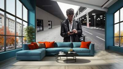 American young man in leather black jacket with backpack with hairstyle with modern mobile phone stands in the city. Handsome hipster guy in trendy clothes stands and looks at the smartphone outdoors. Wall mural