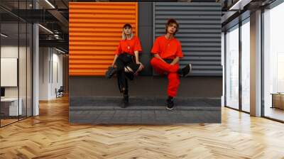 Beautiful fashionable young stylish couple in fashion orange clothes with caps sit near the metal gray and orange wall Wall mural