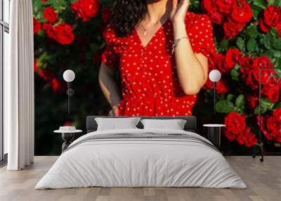 Beautiful woman with fashionable sunglasses and a silver jewelry bracelet in a red dress posing near a bush with red roses Wall mural