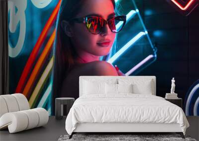 Cool modern urban portrait of a beautiful fashion woman with trendy sunglasses wearing a black top on a background of bright colored neon lights on a dark background Wall mural